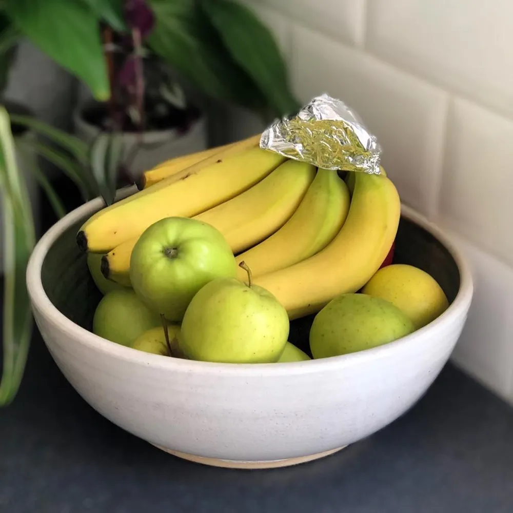 A white fruit bowl with apples and bananas in. The bunch of bananas are wrapped in tinfoil at the stalk.